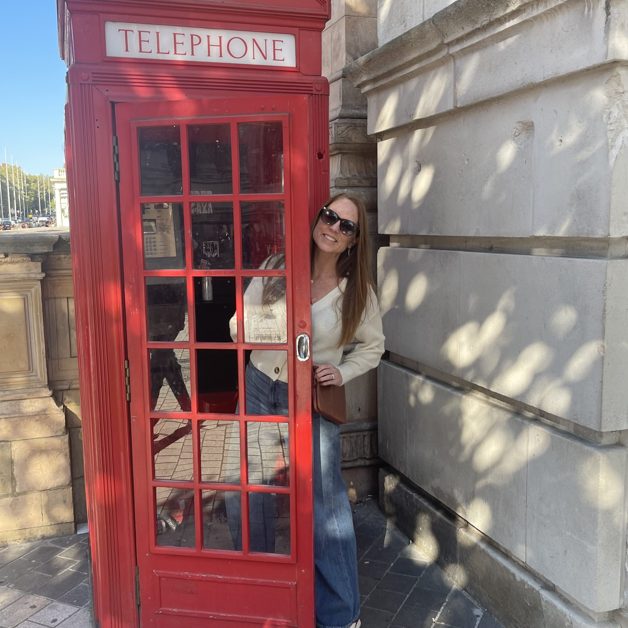 A white woman in a fuzzy white cardigan, sunglasses, and long brown hair leans out of a red phone booth.